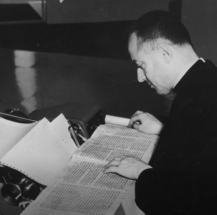 Un sacerdote revisando un libro antiguo y copiando su contenido en un computador de los años 50